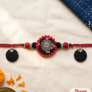 Maroon Chhapa fabric choker with a black printed disc and red pom-pom edge laid flat on handloom with marigolds and brass, beside a pair of round fabric earrings