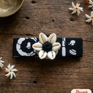 Rongin hair clip in black batik-print cotton with six white cowrie shells around a black thread ball, on a teak table