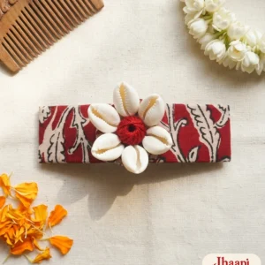Rongin hair clip in red kalamkari-print cotton with a flower of six white cowrie shells around a red thread ball, on a cream handloom cloth